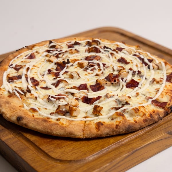 Large cheesy pizza topped with bacon, chicken, mushrooms, and white sauce on a wooden serving board.