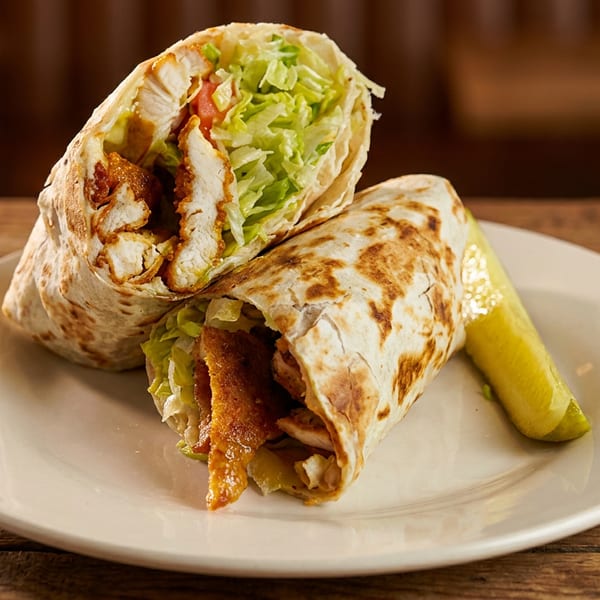 A chicken wrap, cut in half, filled with lettuce and crispy chicken, served with a pickle.