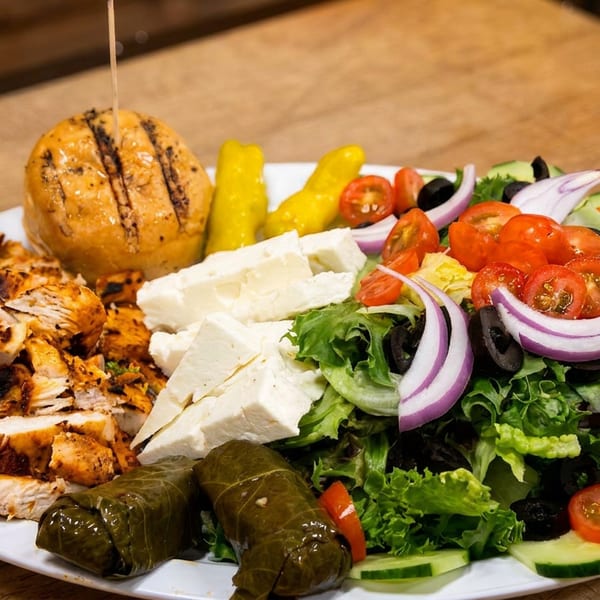 Plate with grilled chicken, Greek salad, feta, stuffed grape leaves, and a grilled bun.