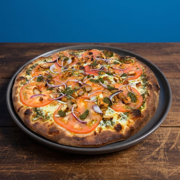 A whole pizza with chicken, tomatoes, red onions, and pesto on a dark plate.