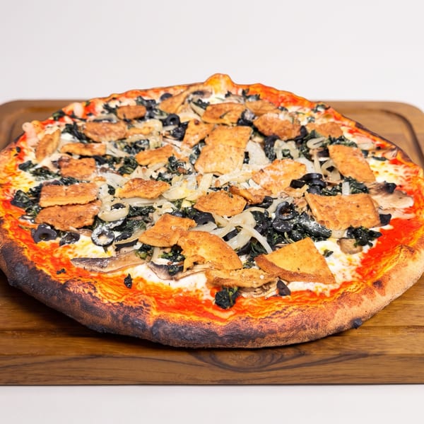 Large round pizza topped with mushrooms, black olives, onions, leafy greens, and crispy pieces on a wooden serving board.