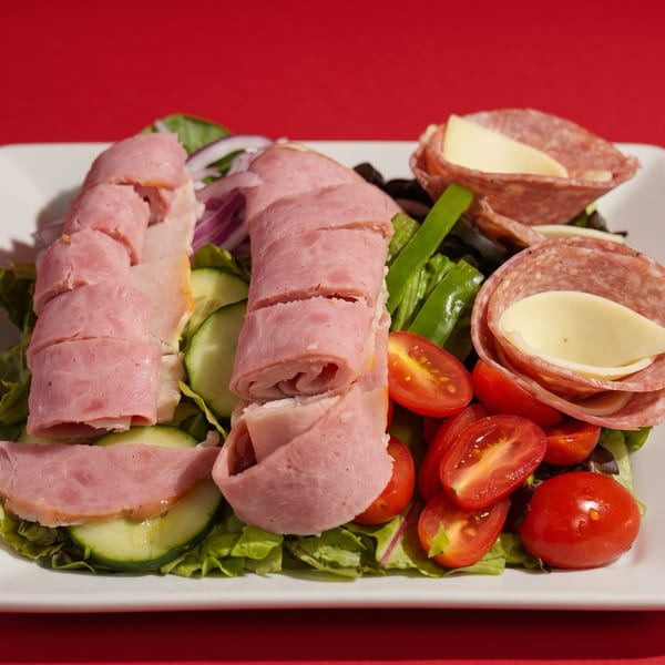 A white plate holds a salad with rolled ham, salami, cheese, tomatoes, cucumbers, and lettuce.