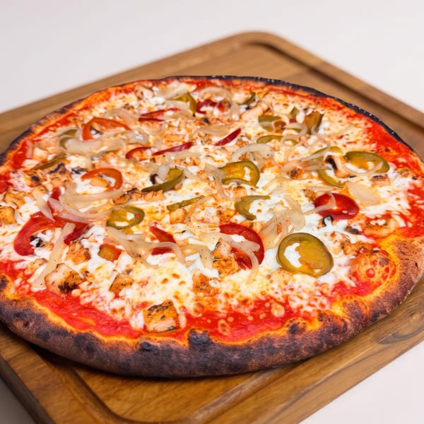 Large wood-fired pizza topped with melted cheese, grilled chicken, onions, peppers, tomatoes, and jalapeños on a wooden board.