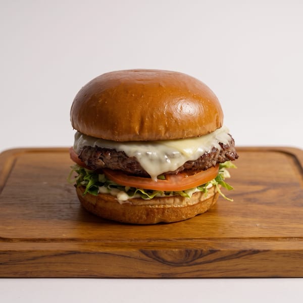 Cheeseburger with a glossy brioche bun, lettuce, tomato, sauce, and melted cheese on a wooden serving board.