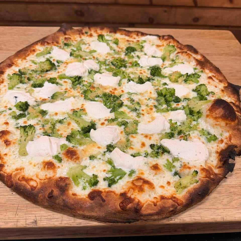 Cheesy pizza topped with broccoli and dollops of white cheese on a wooden serving board.